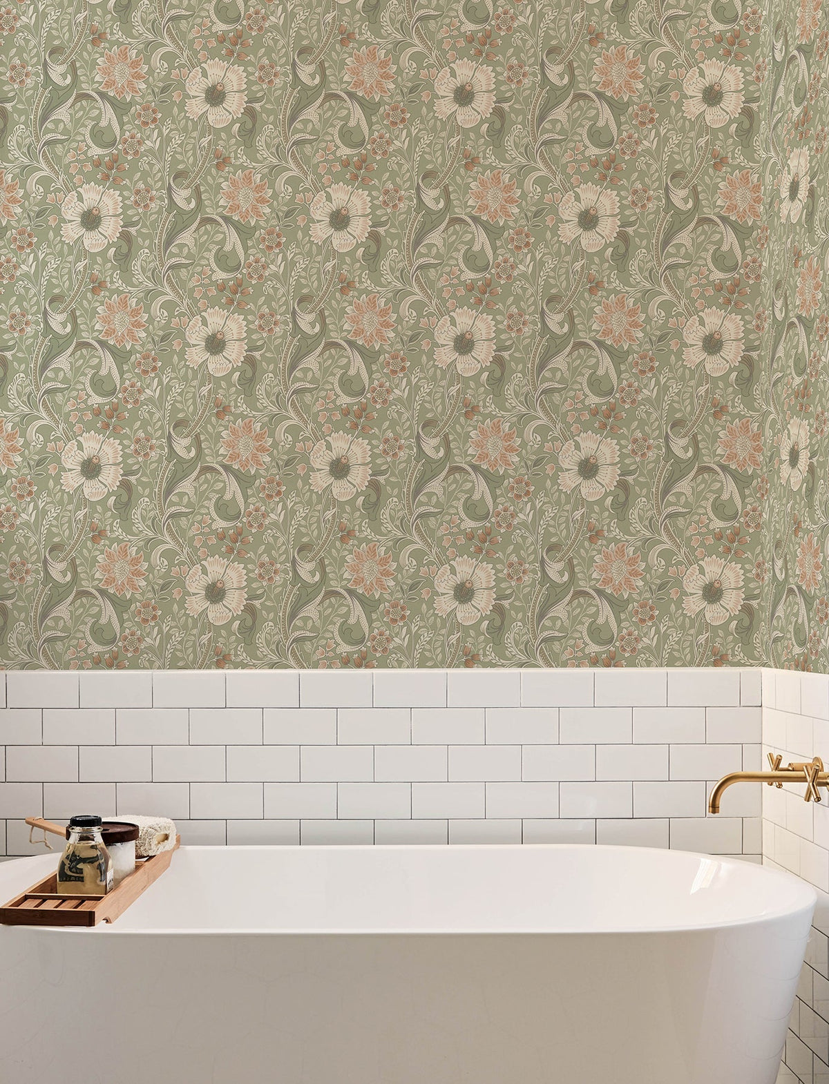 A white bathtub with a gold faucet sits against a wall adorned with Chesapeake Liora Floral Trail Wallpaper by York Wallcoverings, featuring green, pink, and cream floral patterns set above classic white subway tiles. A wooden tray holding soap and bottles rests on the edge of the tub.