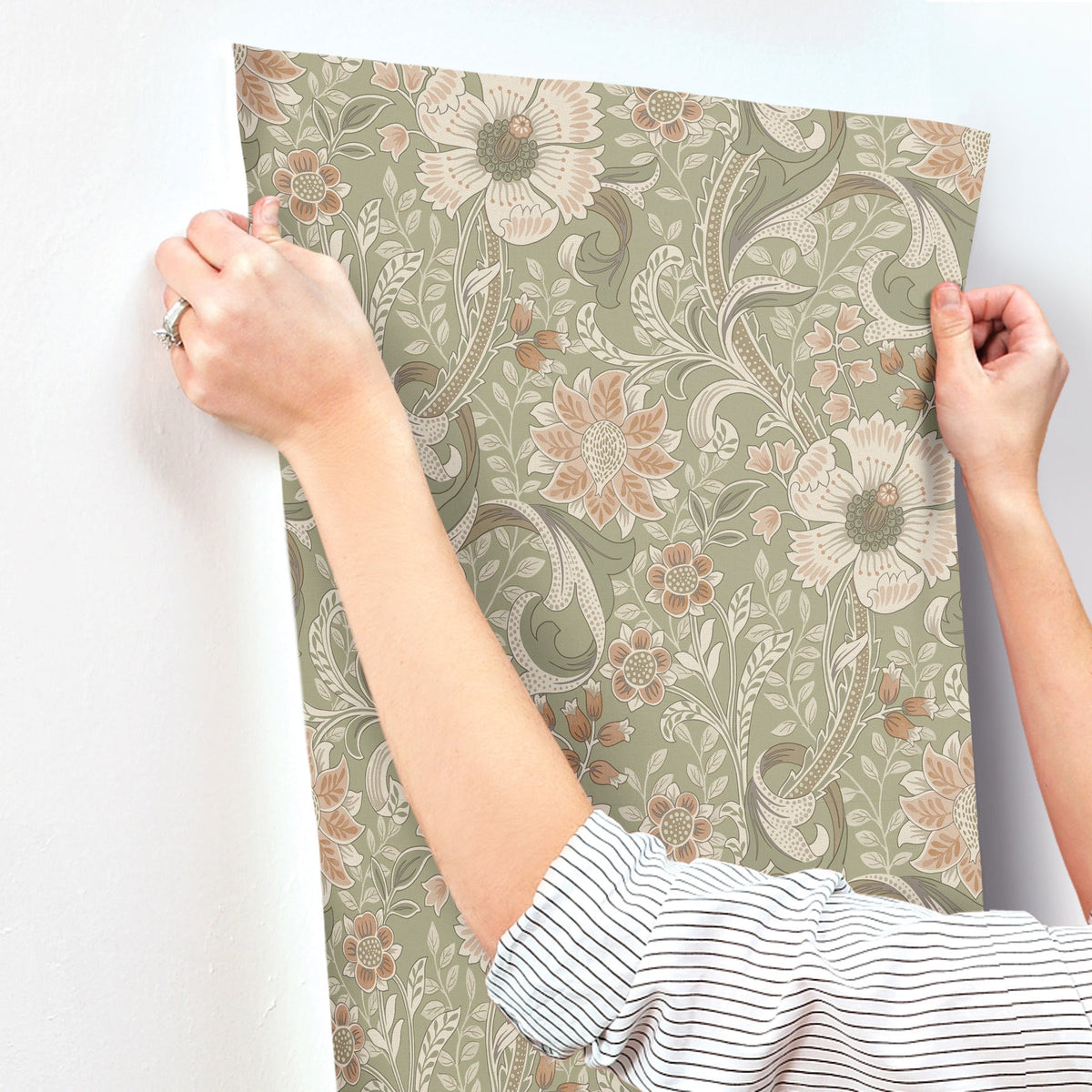 Hands hold up the Chesapeake Liora Floral Trail Wallpaper by York Wallcoverings against a white wall. The wallpaper displays large flowers and leaves in beige, peach, and green tones. The person is wearing a striped shirt and has a ring on their left hand.