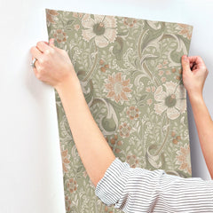 Hands hold up the Chesapeake Liora Floral Trail Wallpaper by York Wallcoverings against a white wall. The wallpaper displays large flowers and leaves in beige, peach, and green tones. The person is wearing a striped shirt and has a ring on their left hand.