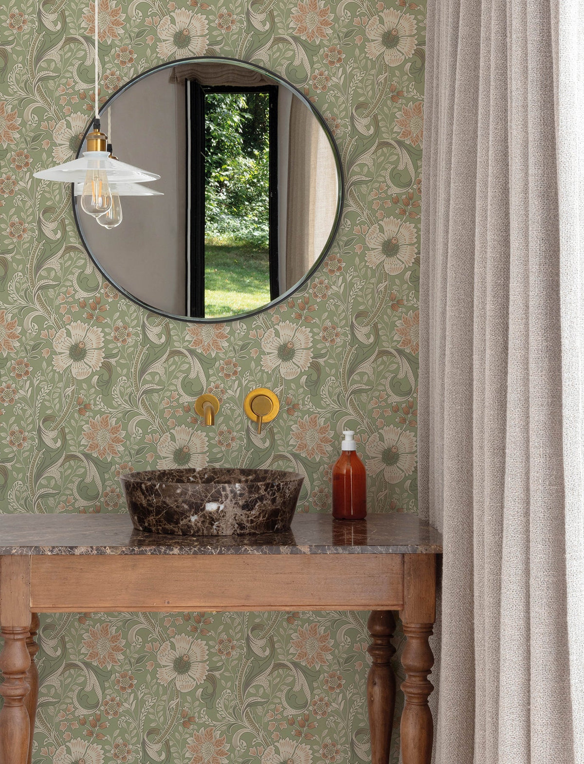 A round mirror hangs above a marble sink with gold fixtures on a wooden vanity. The York Wallcoverings Chesapeake Liora Floral Trail Wallpaper decorates the walls, complemented by beige curtains. A soap dispenser and pendant light are also present, while a window reveals greenery outside.