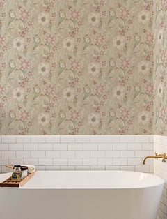A bathtub with a gold faucet is set against a wall featuring white subway tiles and Chesapeake Liora Floral Trail Wallpaper by York Wallcoverings in cream, pink, and green. A wooden tray holds bath items on the tub’s edge.