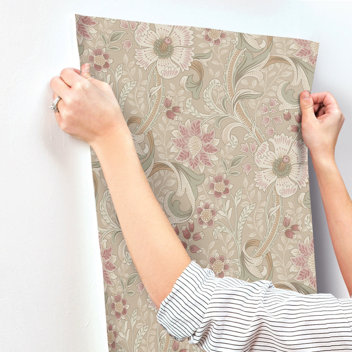 Someone with rolled-up sleeves applies the Chesapeake Liora Floral Trail Wallpaper by York Wallcoverings, featuring pink and cream flowers with green leaves, onto a white wall. Only their hands, arms, and torso are shown.
