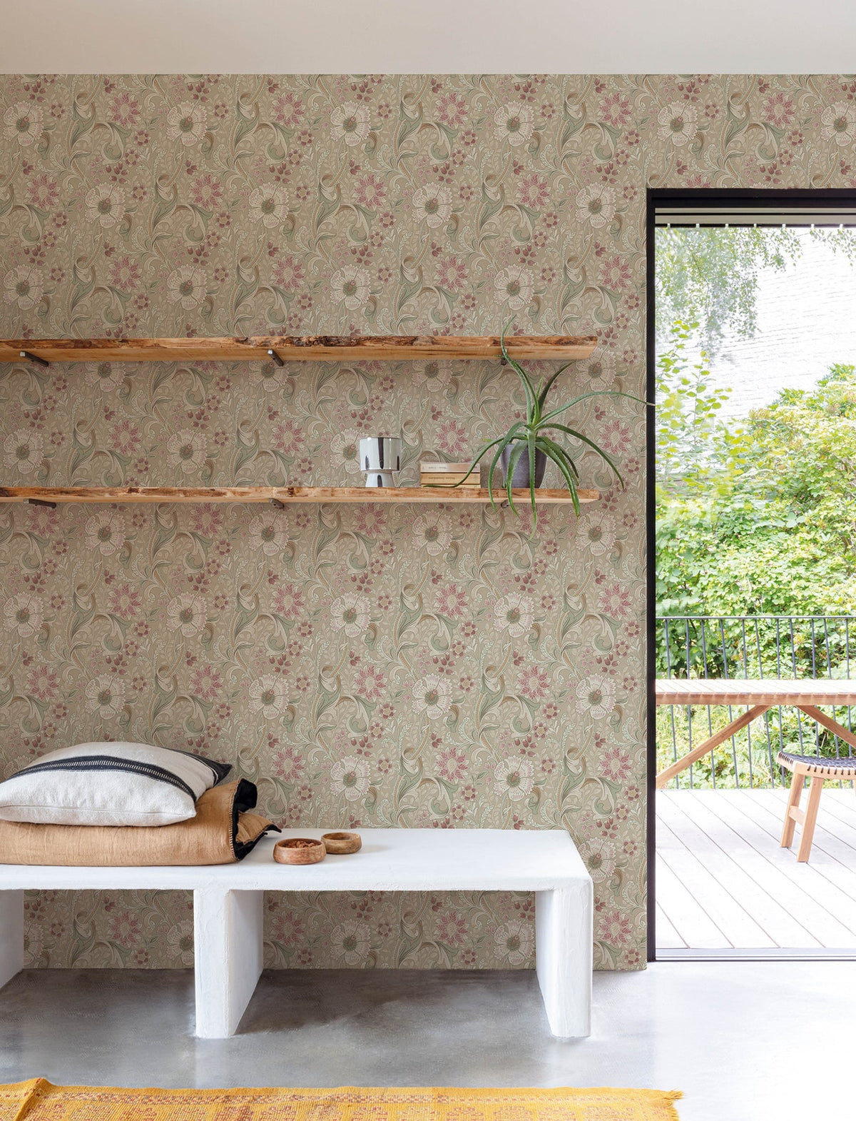 A minimal room featuring York Wallcoverings’ Chesapeake Liora Floral Trail Wallpaper, with wooden shelves holding books and a plant, a white bench with cushions, and a view of a green garden through an open glass door.
