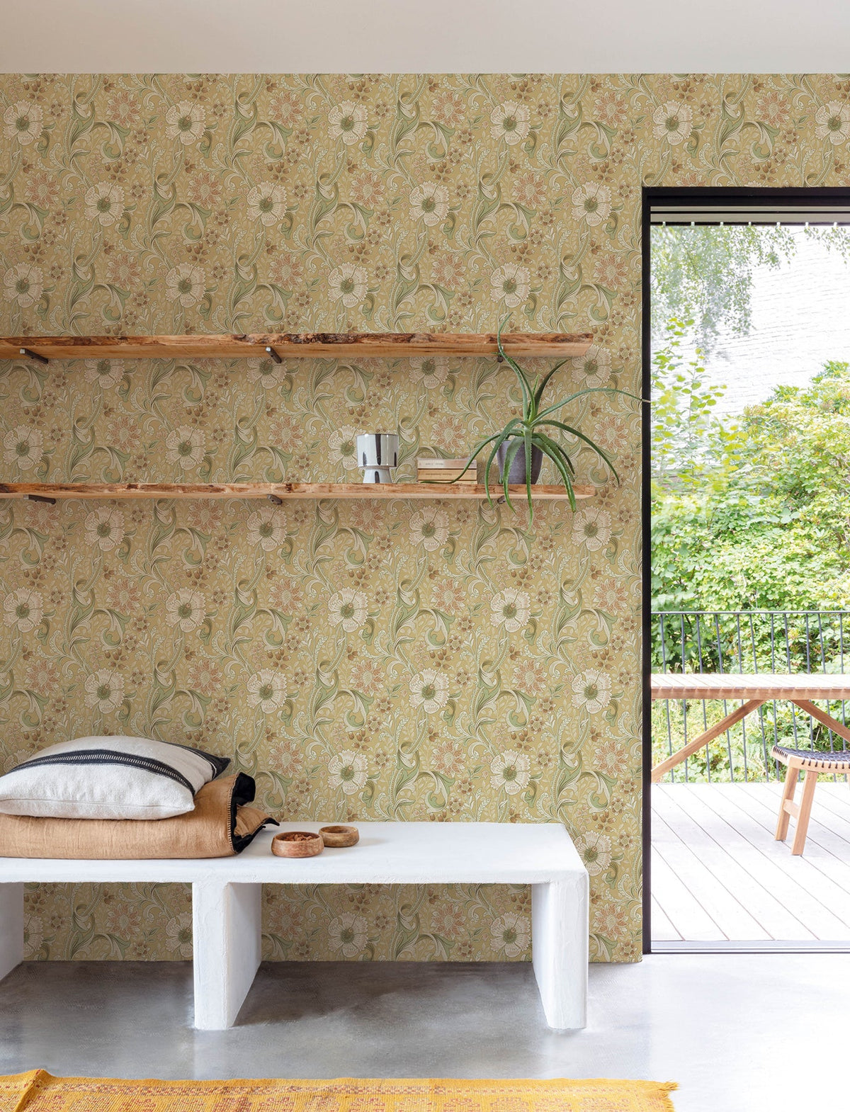 A room featuring Chesapeake Liora Floral Trail Wallpaper by York Wallcoverings, with wooden shelves displaying a plant and decor accents, and a white bench topped with cushions; a door leads out to a patio where trees and outdoor furniture are visible.