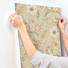 A person is installing Chesapeake Liora Floral Trail Wallpaper by York Wallcoverings onto a white wall. Only their hands and arms, dressed in a striped long-sleeve shirt and adorned with a ring, are visible.