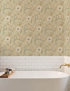 A white bathtub with a wooden tray sits against a wall adorned with Chesapeake Liora Floral Trail Wallpaper by York Wallcoverings and white subway tiles, featuring gold fixtures.