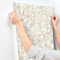 Wearing a striped shirt and a ring, a person applies York Wallcoverings’ Chesapeake Laverne Wildflowers Wallpaper—featuring a delicate floral pattern in soft beige, blue, and yellow tones—to a white wall; only their hands and arms are visible.