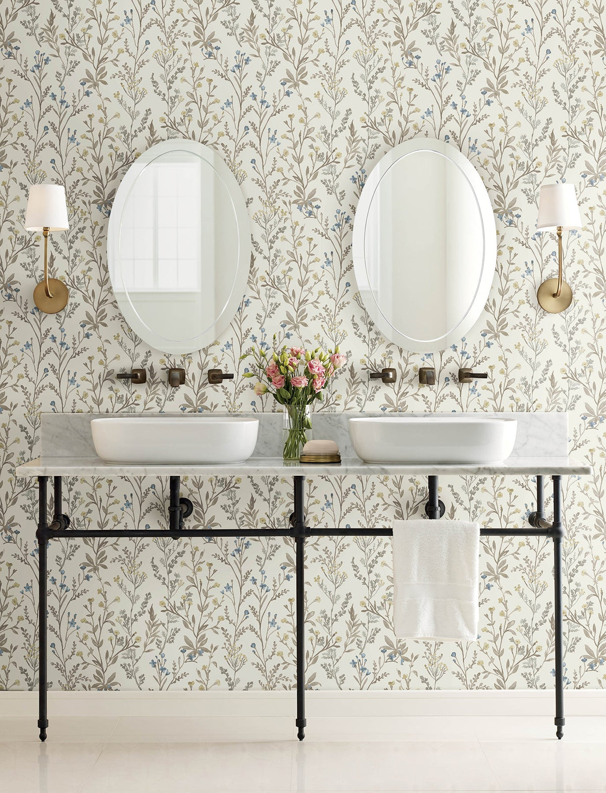 Two sinks with oval mirrors above each, brass sconces on both sides, Chesapeake Laverne Wildflowers Wallpaper by York Wallcoverings adorning the walls, a vase of flowers between the sinks, and a white towel hanging under the marble countertop.