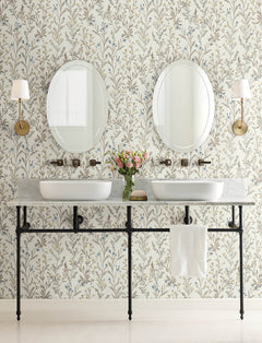 Two sinks with oval mirrors above each, brass sconces on both sides, Chesapeake Laverne Wildflowers Wallpaper by York Wallcoverings adorning the walls, a vase of flowers between the sinks, and a white towel hanging under the marble countertop.