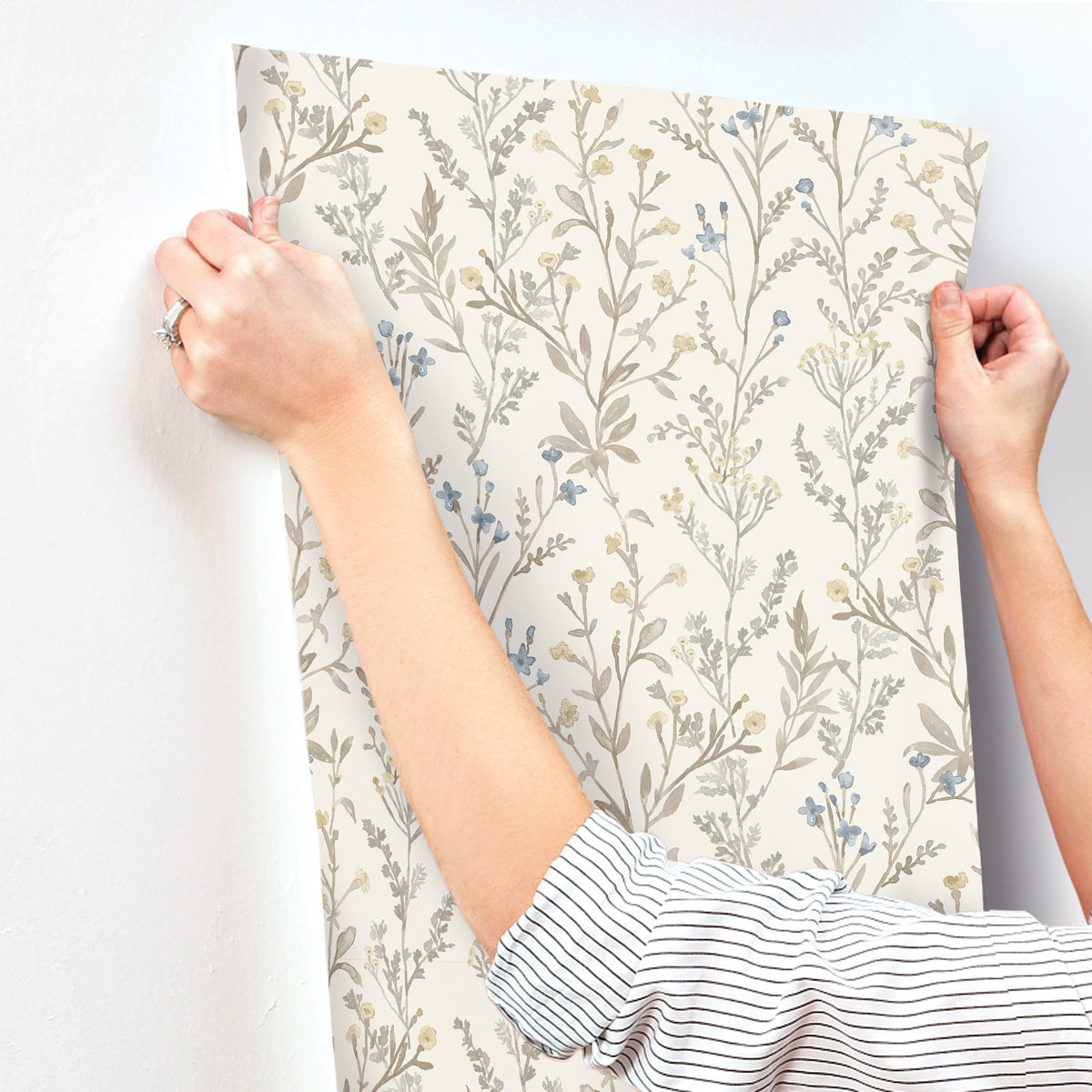 A person wearing a striped shirt and a ring on their left hand is applying Chesapeake Laverne Wildflowers Wallpaper by York Wallcoverings, featuring beige, blue, and yellow small flowers and leaves, to a white wall. Only their arms and hands are visible.