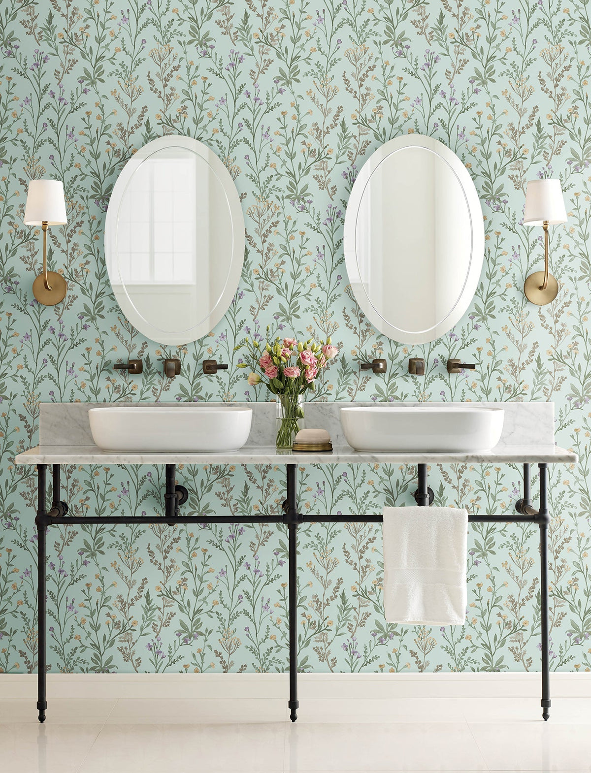 Double sink bathroom vanity with a marble countertop, two round mirrors, wall-mounted faucets, brass sconces, a white towel, and a vase of flowers set against York Wallcoverings’ Chesapeake Laverne Wildflowers Wallpaper.