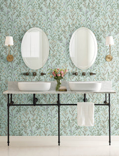 Double sink bathroom vanity with a marble countertop, two round mirrors, wall-mounted faucets, brass sconces, a white towel, and a vase of flowers set against York Wallcoverings’ Chesapeake Laverne Wildflowers Wallpaper.