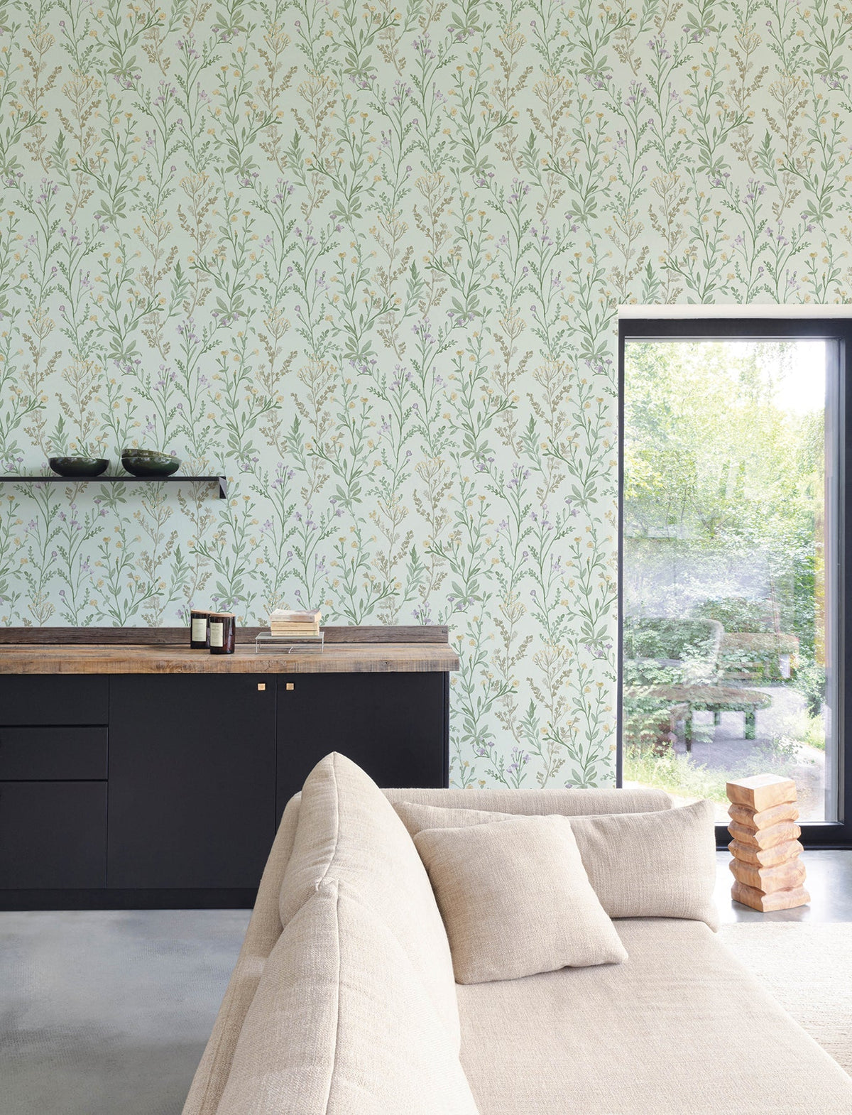 A modern living room featuring a beige sofa, black cabinets, wooden countertop, and Chesapeake Laverne Wildflowers Wallpaper by York Wallcoverings. A window reveals a lush garden outside, while Jenga blocks are stacked on the floor near the sofa.