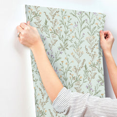 A person applies York Wallcoverings’ Chesapeake Laverne Wildflowers Wallpaper to a white wall. Only their arms and hands are visible; they wear a striped shirt and a ring. The wallpaper displays green stems, leaves, and small flowers.