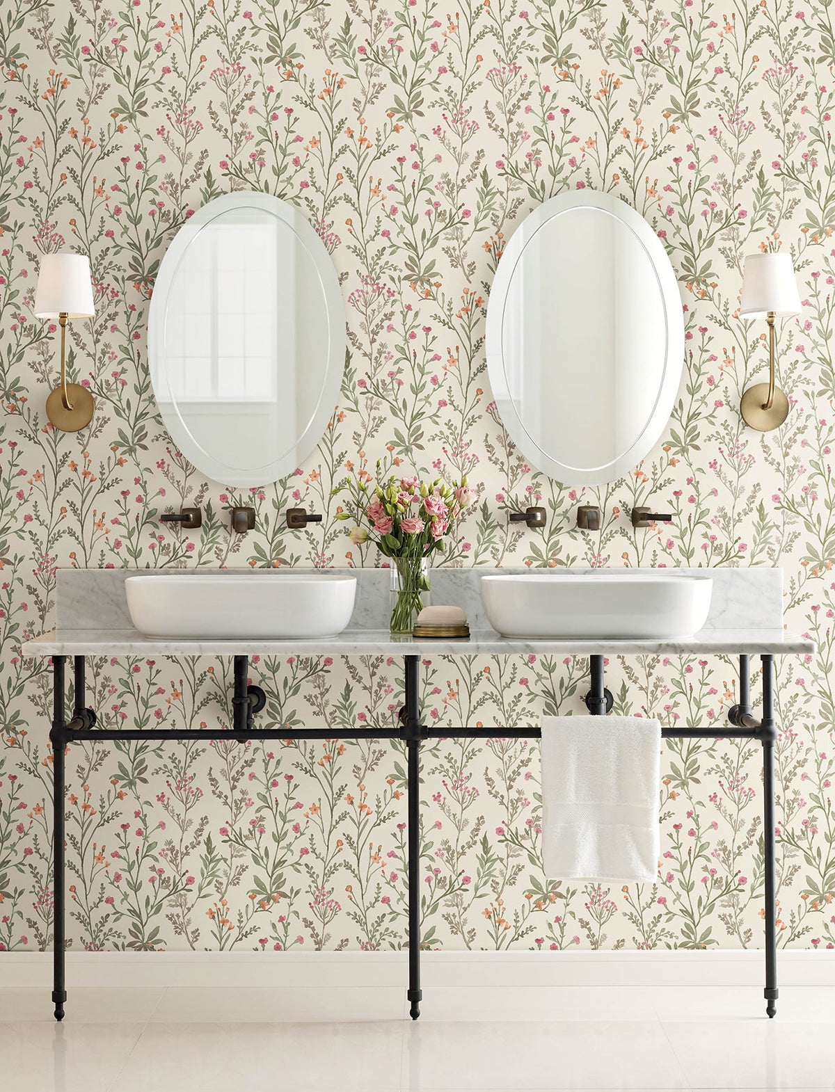 A bathroom featuring York Wallcoverings’ Chesapeake Laverne Wildflowers Wallpaper, two round mirrors above dual sinks, wall sconces, a shared marble countertop with a towel hanging on a metal rail, and a vase of flowers placed in the center.