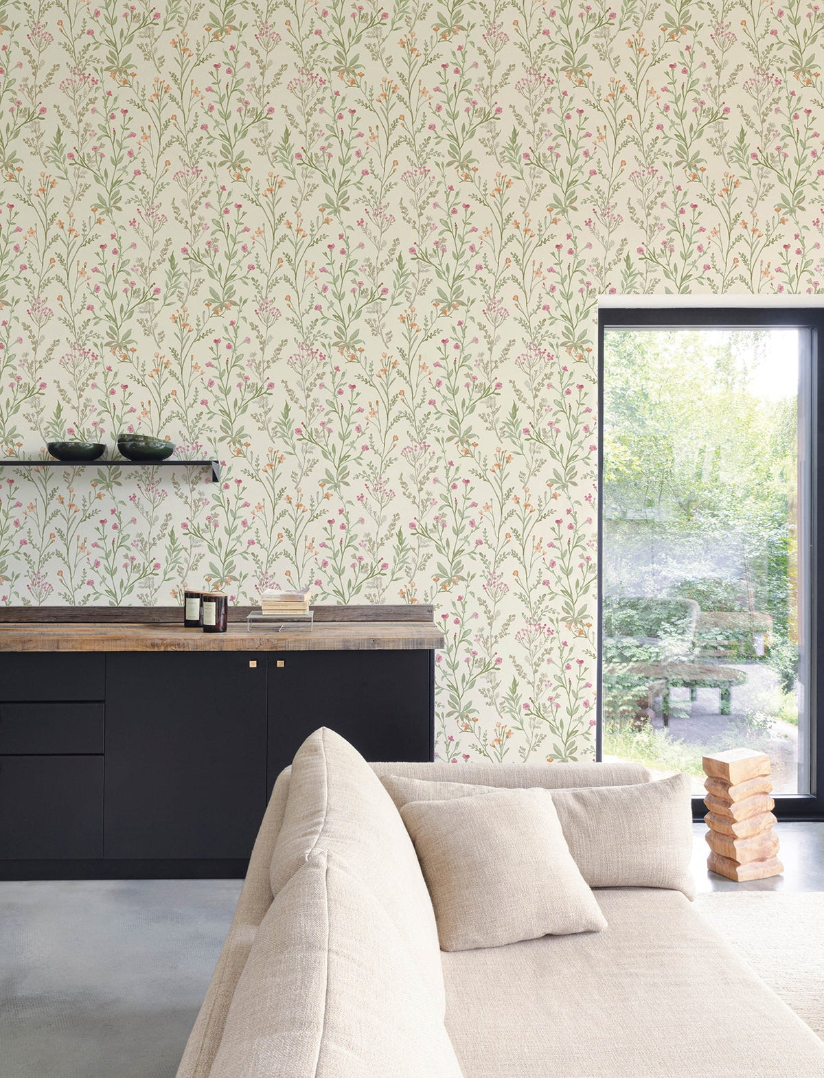 A modern living room features a beige sofa, black cabinetry, and a wooden countertop. Chesapeake Laverne Wildflowers Wallpaper by York Wallcoverings adds a floral touch to the walls. A large window reveals a green garden outside, and a stack of wooden blocks lies on the floor.