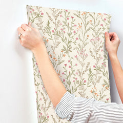 Hands holding up Chesapeake Laverne Wildflowers Wallpaper by York Wallcoverings, featuring a cream background adorned with pink, orange, and green botanical patterns, against a white wall. The person is wearing a striped shirt and a ring on their left hand.
