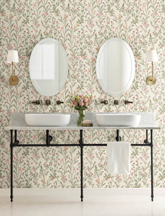 A bathroom featuring Chesapeake Laverne Wildflowers Wallpaper by York Wallcoverings, two round mirrors, wall sconces, twin sinks set in a marble counter, and a vase of flowers in the center. A towel hangs beneath the counter.