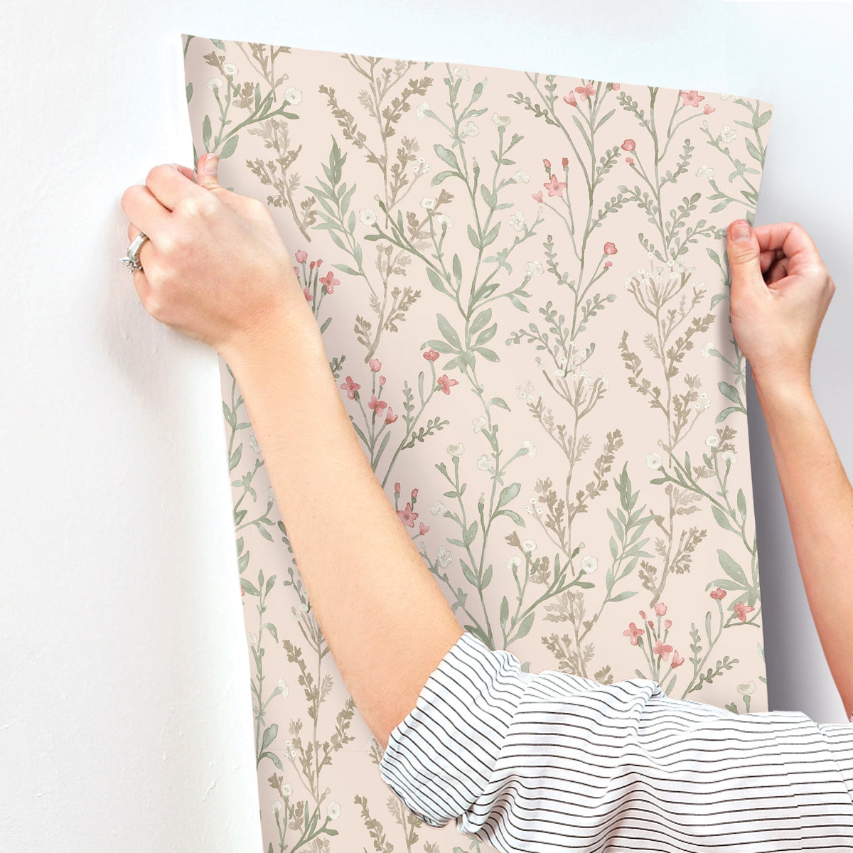 A person in a striped shirt is applying the Chesapeake Laverne Wildflowers Wallpaper by York Wallcoverings, which features pink and green botanical patterns, to a white wall.