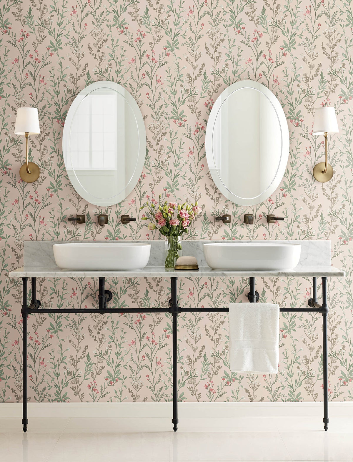 Double bathroom vanity with two vessel sinks, round mirrors, brass sconces, a towel hanging below, and a vase of pink flowers on the counter. The wall is adorned with Chesapeake Laverne Wildflowers Wallpaper by York Wallcoverings in soft pink and green tones.
