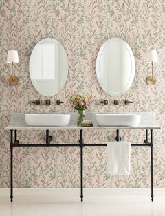 Double bathroom vanity with two vessel sinks, round mirrors, brass sconces, a towel hanging below, and a vase of pink flowers on the counter. The wall is adorned with Chesapeake Laverne Wildflowers Wallpaper by York Wallcoverings in soft pink and green tones.