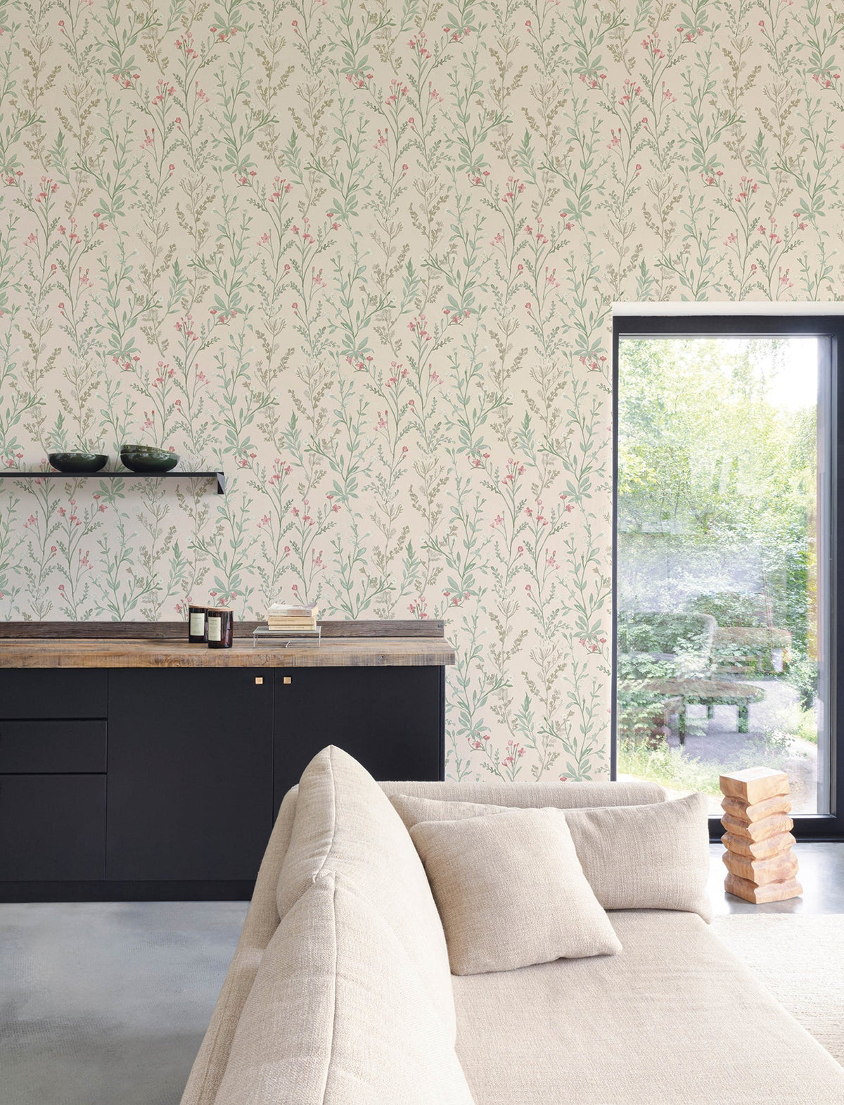 A modern living room featuring York Wallcoverings’ Chesapeake Laverne Wildflowers Wallpaper in beige, black cabinets, a wooden countertop, a floating shelf with bowls, a beige sofa, a Jenga game set, and a large window overlooking a green garden.