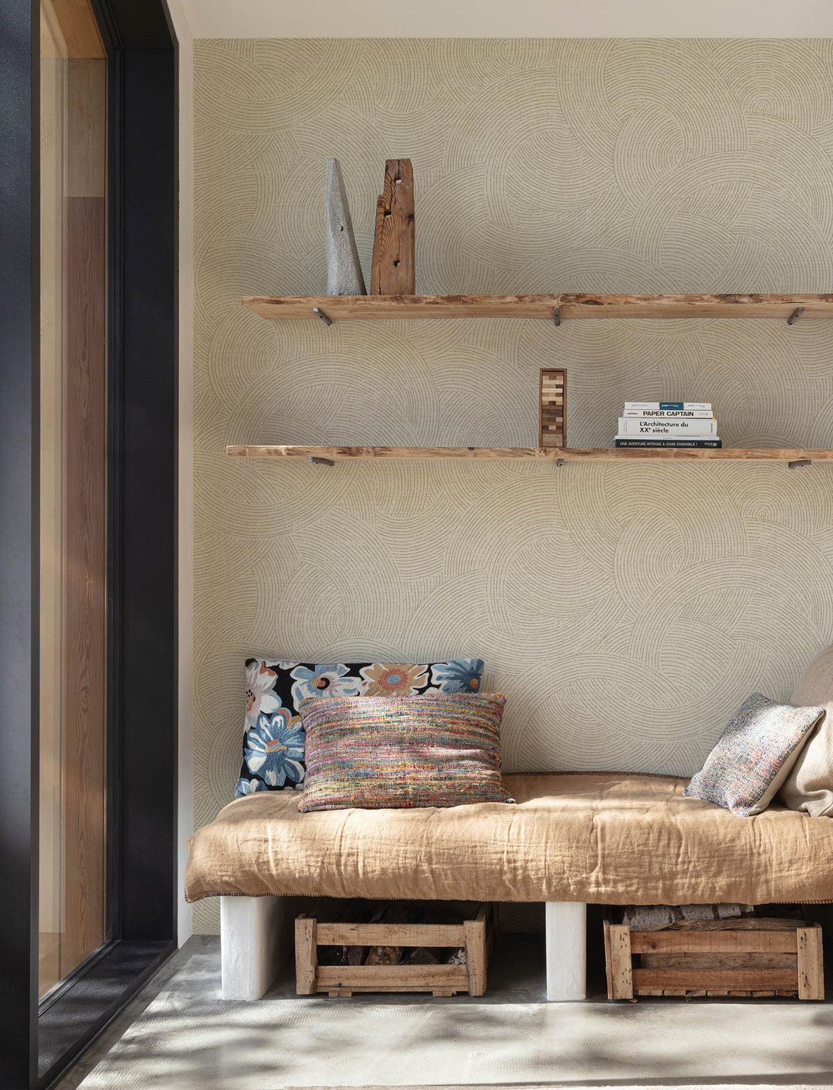 The cozy nook features York Wallcoverings’ Chesapeake Harrison Swirling Geometric Wallpaper, with a tan blanket, colorful pillows, wooden shelves displaying books and decor, and two wooden crates placed underneath—all beside a large window that fills the space with natural light.