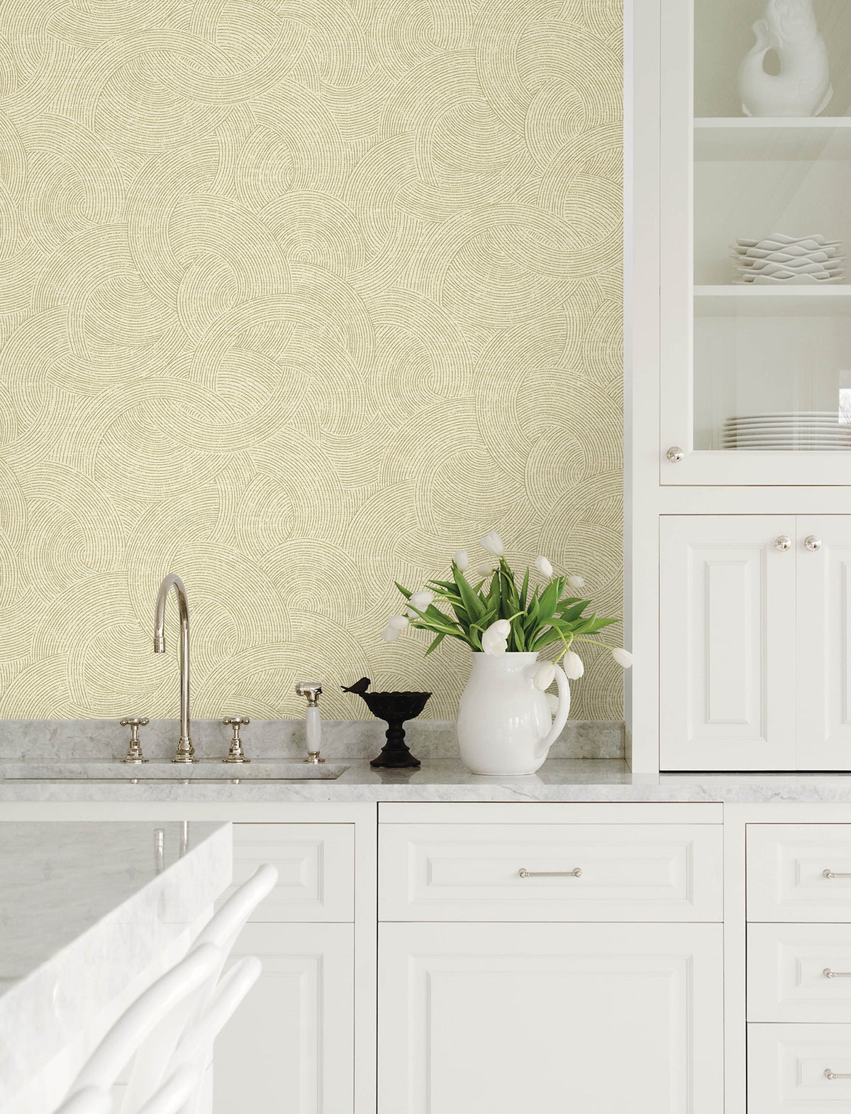 Modern kitchen with white cabinets, marble countertops, a sink with a faucet, a vase of white tulips, a decorative black bird figure, and an open cabinet displaying white dishes set against walls adorned with Chesapeake Harrison Swirling Geometric Wallpaper by York Wallcoverings.