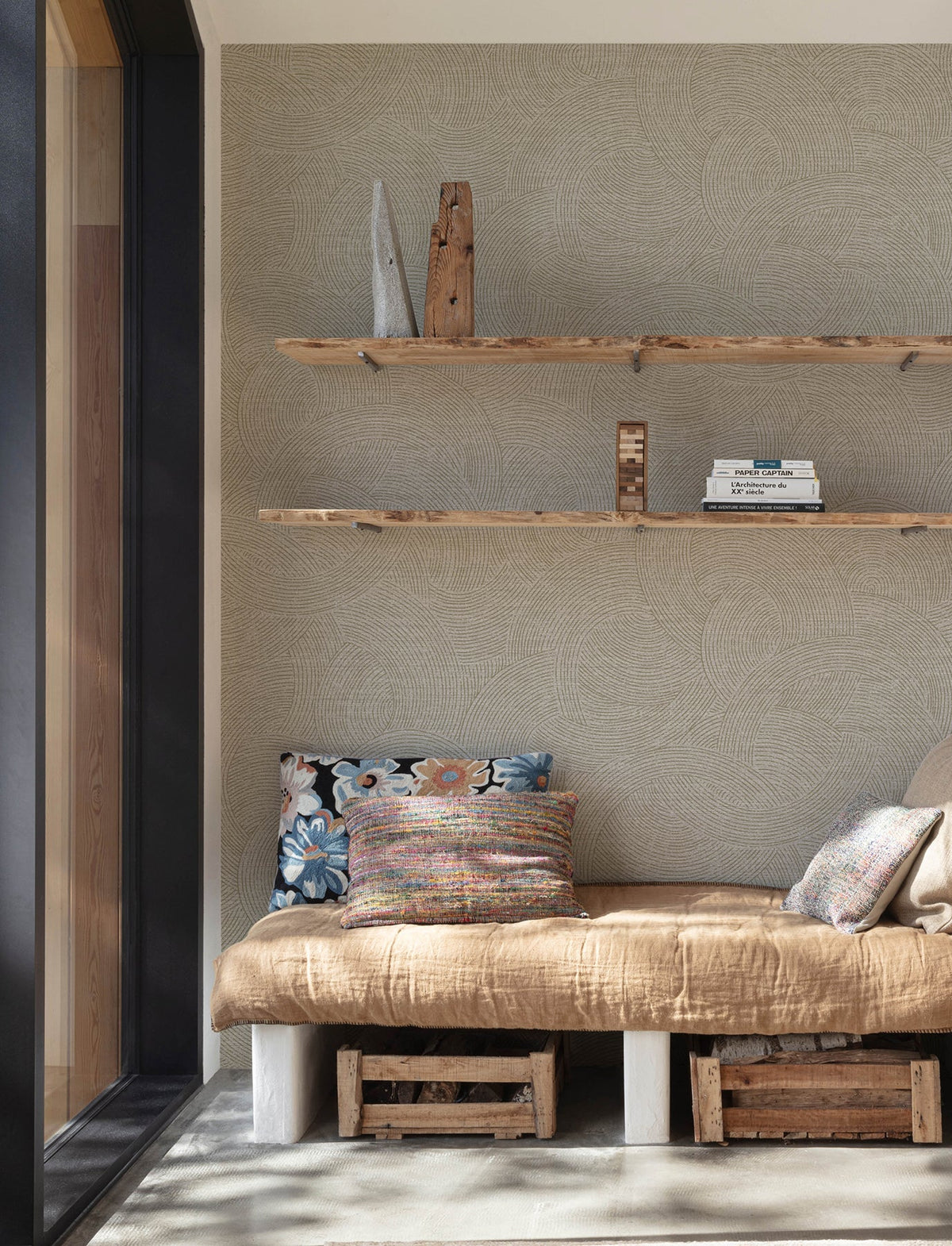 A cozy reading nook featuring a beige cushion, patterned pillows, and wooden shelves displaying books and decor is set against Chesapeake Harrison Swirling Geometric Wallpaper by York Wallcoverings. Wooden crates are neatly stored underneath the seat.