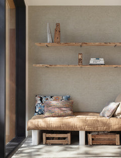 Create a cozy corner featuring Chesapeake Harrison Swirling Geometric Wallpaper by York Wallcoverings, a built-in bench topped with a tan blanket and colorful pillows. Wooden shelves above display books and decor, while storage crates sit below the bench. Sunlight pours in through a large window on the left, illuminating the inviting space.