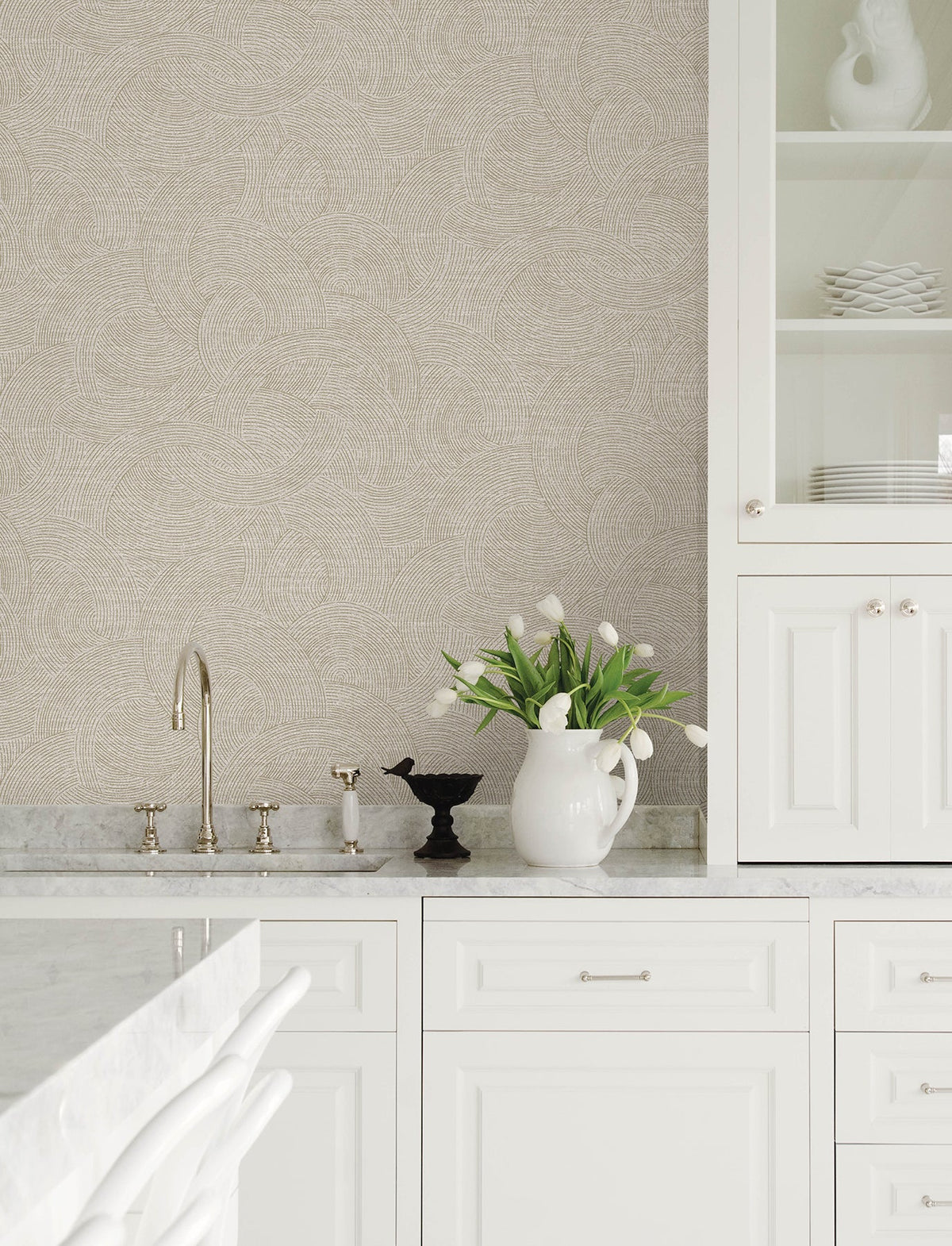 Bright, modern kitchen featuring white cabinets, marble countertops, a gold faucet, and a white pitcher of tulips. Chesapeake Harrison Swirling Geometric Wallpaper by York Wallcoverings adds stylish flair to the space, while neatly stacked white dishes are displayed in a glass cabinet.