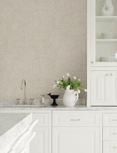Bright, modern kitchen featuring white cabinets, marble countertops, a gold faucet, and a white pitcher of tulips. Chesapeake Harrison Swirling Geometric Wallpaper by York Wallcoverings adds stylish flair to the space, while neatly stacked white dishes are displayed in a glass cabinet.