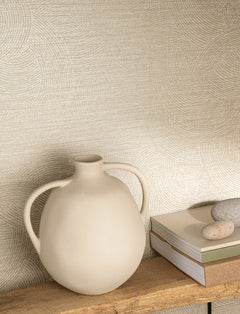 A round, cream-colored vase with two handles sits on a wooden shelf next to stacked books and smooth stones, set against a wall adorned with Chesapeake Harrison Swirling Geometric Wallpaper by York Wallcoverings.