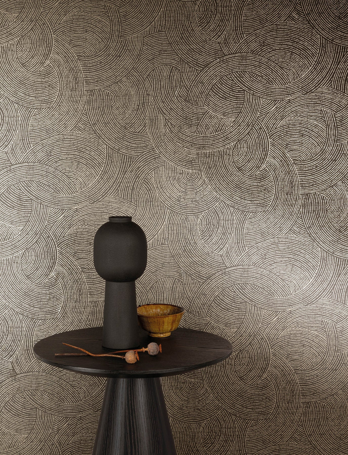 A round dark table with a tall, matte black vase, a small yellow bowl, and a twig sits in front of a beige wall adorned with York Wallcoverings’ Chesapeake Harrison Swirling Geometric Wallpaper.