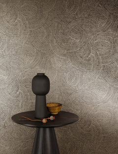 A round dark table with a tall, matte black vase, a small yellow bowl, and a twig sits in front of a beige wall adorned with York Wallcoverings’ Chesapeake Harrison Swirling Geometric Wallpaper.