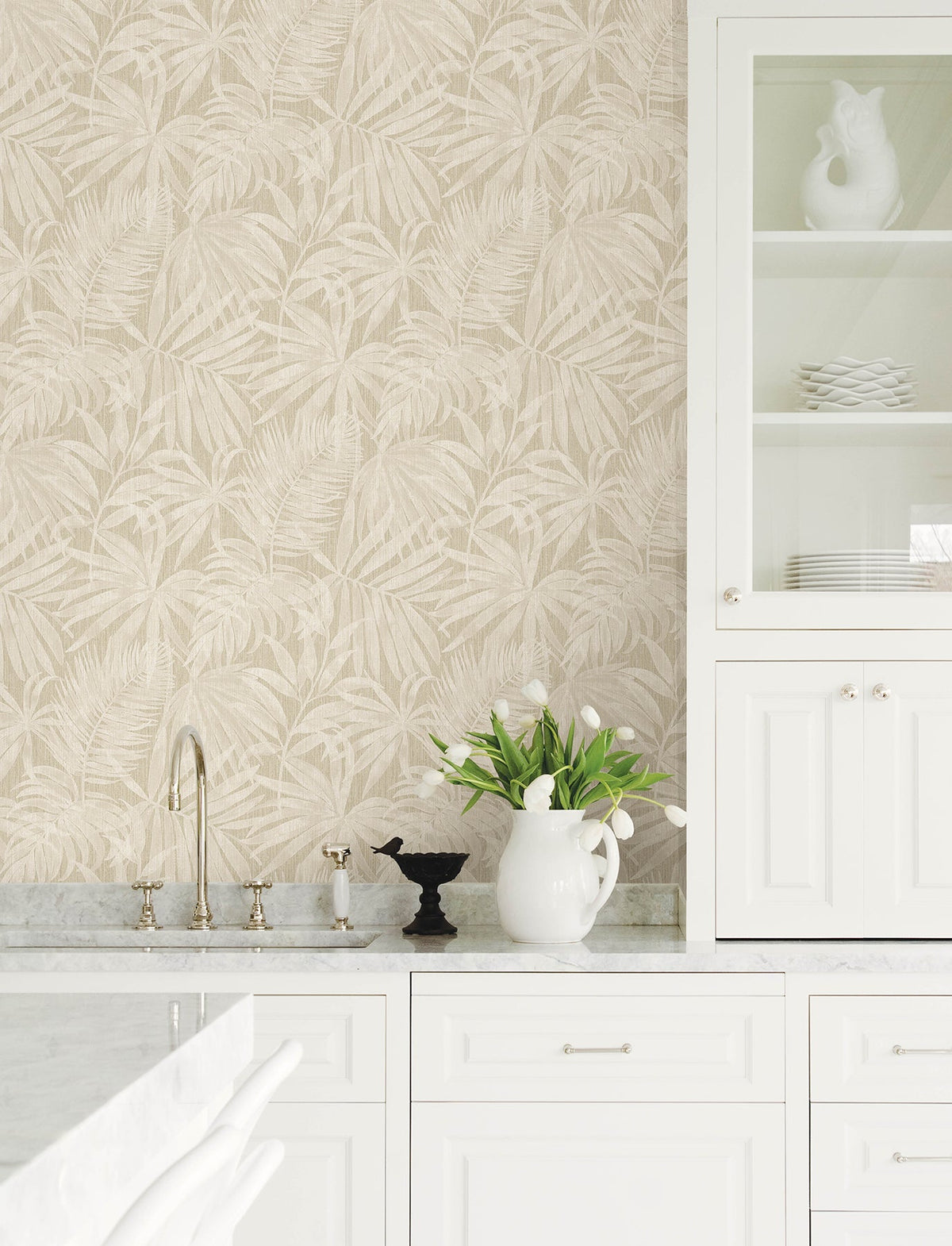 Bright white kitchen with a marble countertop, Chesapeake Sonny Fronds Wallpaper by York Wallcoverings, a sink, and open cabinets with dishes. A white pitcher filled with tulips and a black decorative cup sit on the counter.