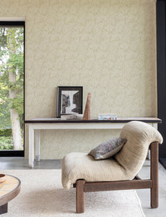 A modern lounge chair with a textured cushion sits in front of a narrow console table adorned with framed art, a wooden sculpture, and a box, set against a wall covered in Chesapeake Sonny Fronds Wallpaper by York Wallcoverings, near a window that reveals lush green trees outside.