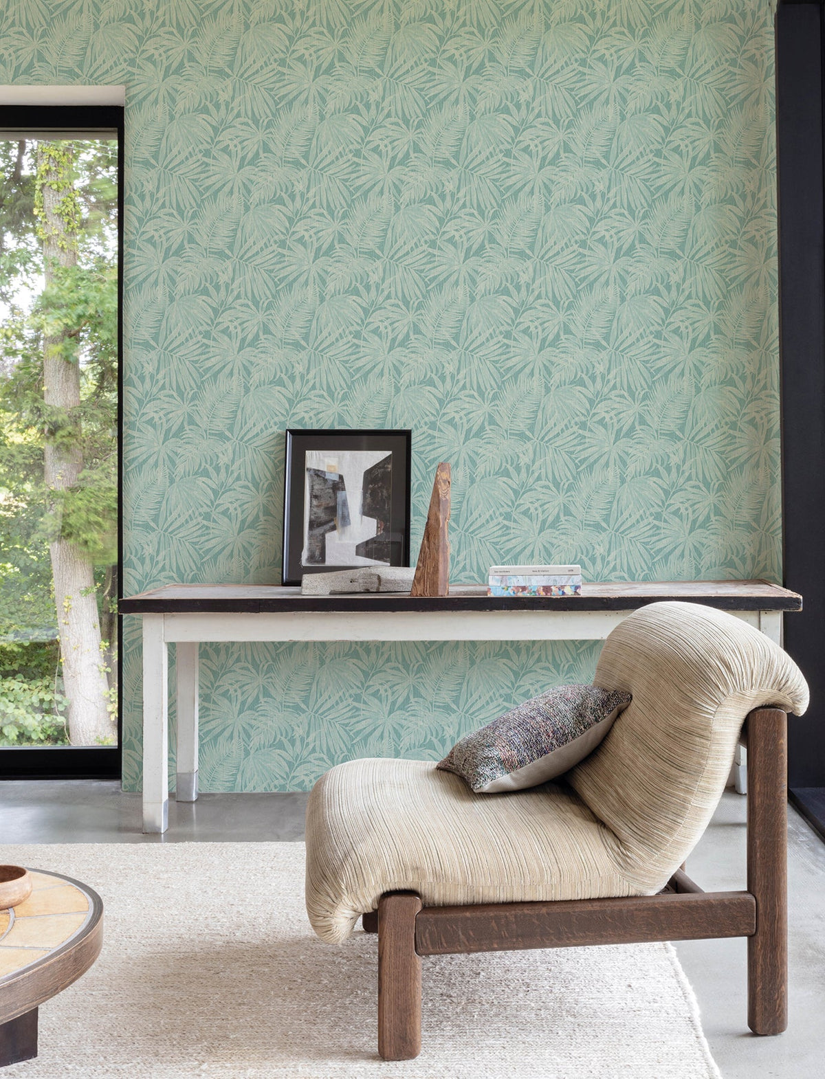 A modern living room features a textured beige chair, a slim console table adorned with framed art and decor, and walls covered in York Wallcoverings’ Chesapeake Sonny Fronds Wallpaper in a soft pastel green. A large window offers views of lush greenery outside.