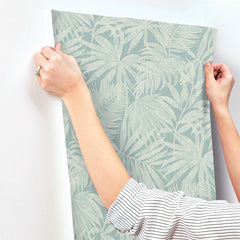 Wearing a striped sleeve, a person smooths a sheet of Chesapeake Sonny Fronds Wallpaper by York Wallcoverings—featuring a pale green leafy pattern—onto a white wall, securing it at the top corners with both hands.