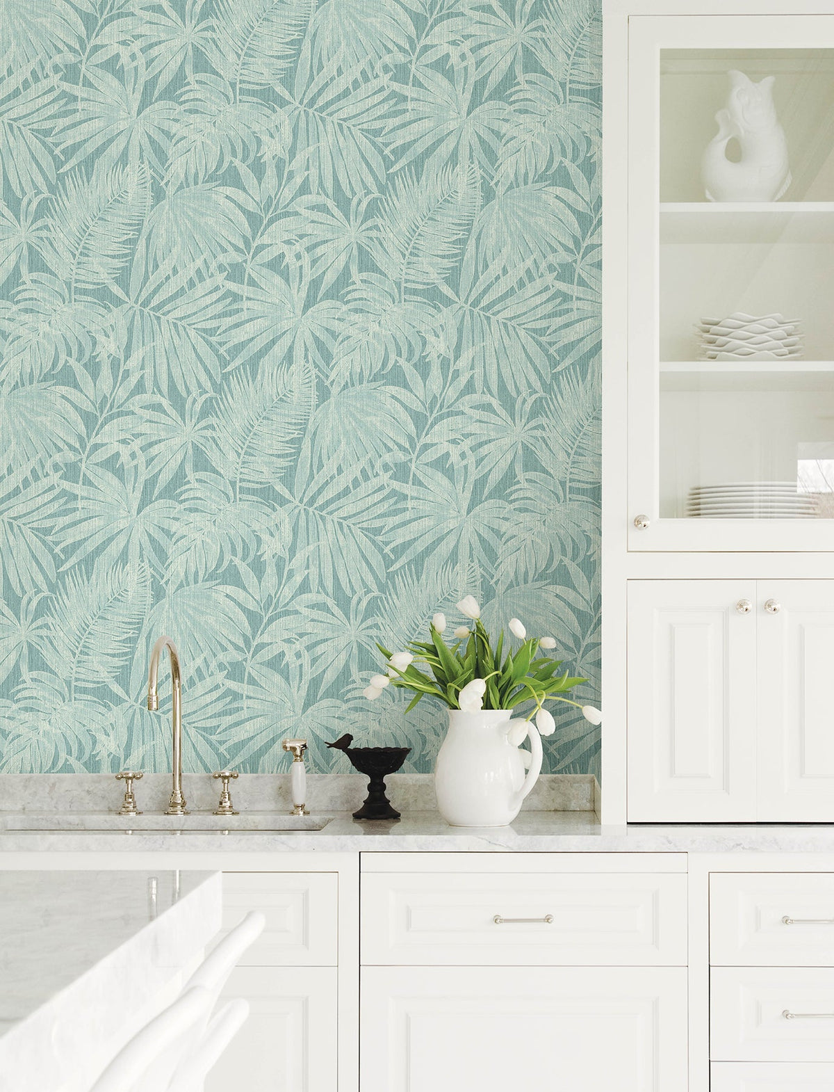White kitchen with marble countertop, gold faucet, and Chesapeake Sonny Fronds Wallpaper by York Wallcoverings in a blue-green palm leaf pattern. White dishes and a pitcher with tulips are displayed on the counter and inside a glass-front cabinet.