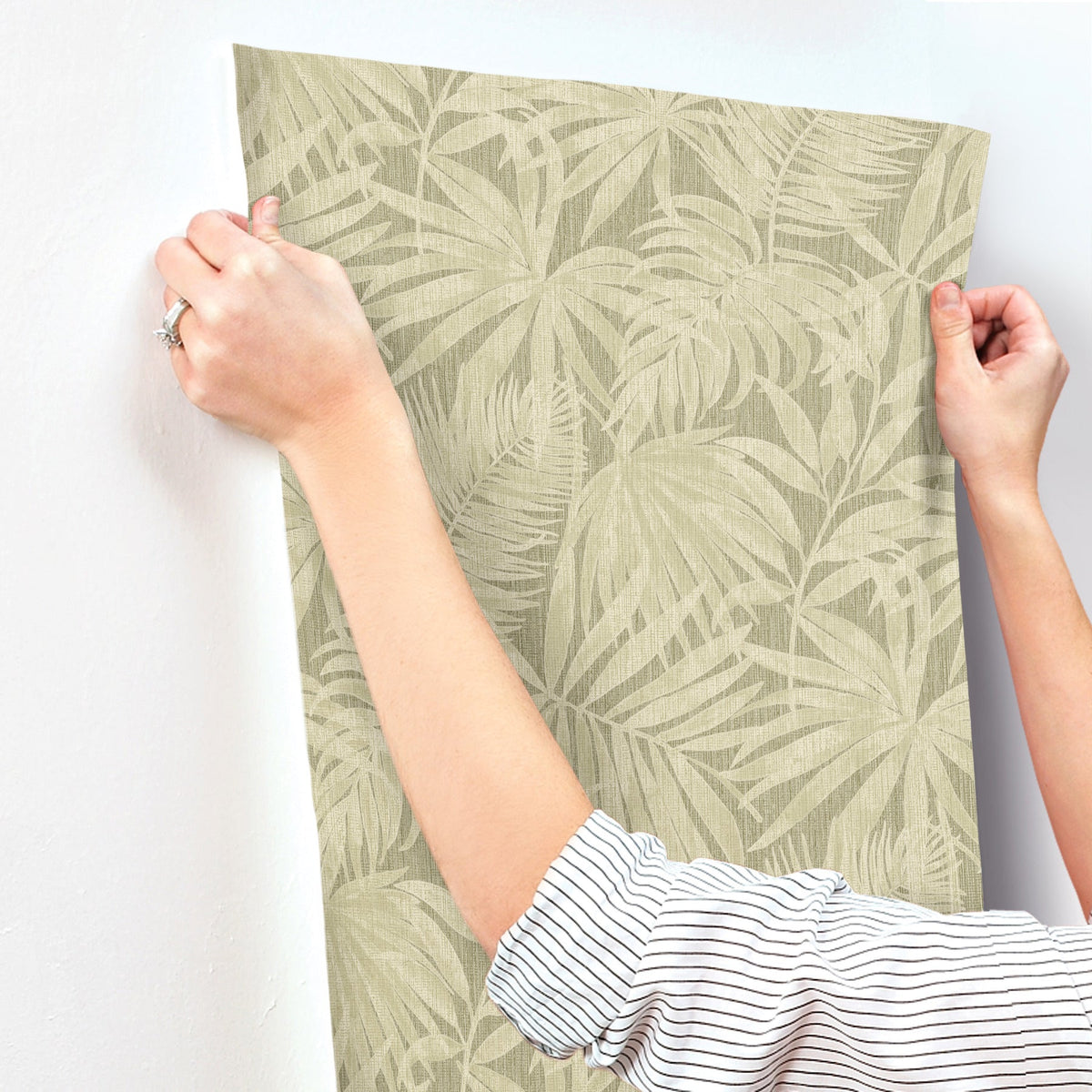 A person in a striped shirt is installing York Wallcoverings Chesapeake Sonny Fronds wallpaper—featuring a beige, leafy pattern—on a white wall, holding the top corners of the wallpaper with both hands.