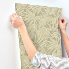 A person in a striped shirt applies Chesapeake Sonny Fronds Wallpaper by York Wallcoverings, featuring a leafy green pattern, to a white wall.