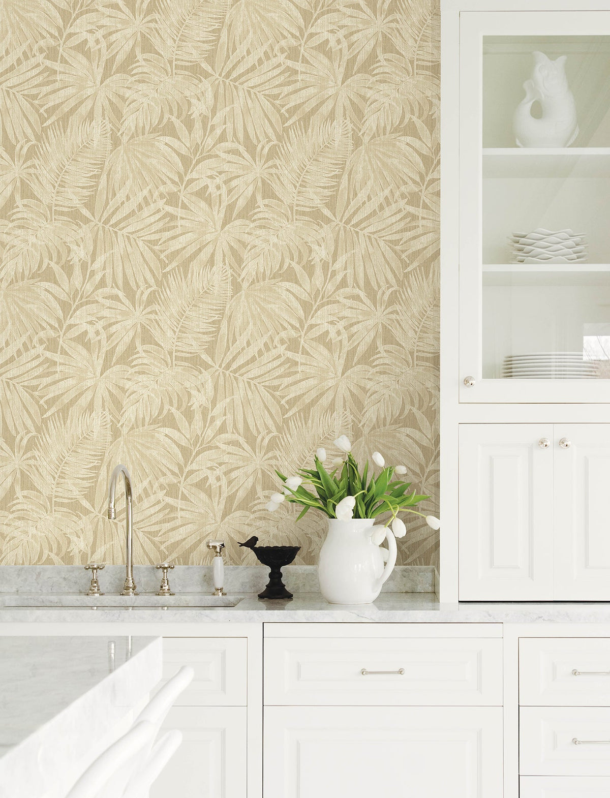 Bright white kitchen with marble countertops, Chesapeake Sonny Fronds Wallpaper by York Wallcoverings in beige, tulips in a white pitcher, black bird decor, and glass-front cabinets holding plates and a white ceramic rooster.