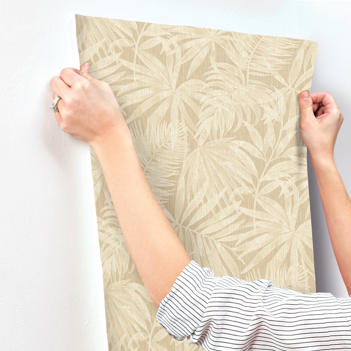 A person with a striped sleeve presses Chesapeake Sonny Fronds Wallpaper by York Wallcoverings—featuring a beige leafy pattern—onto a white wall, smoothing it with their hands.
