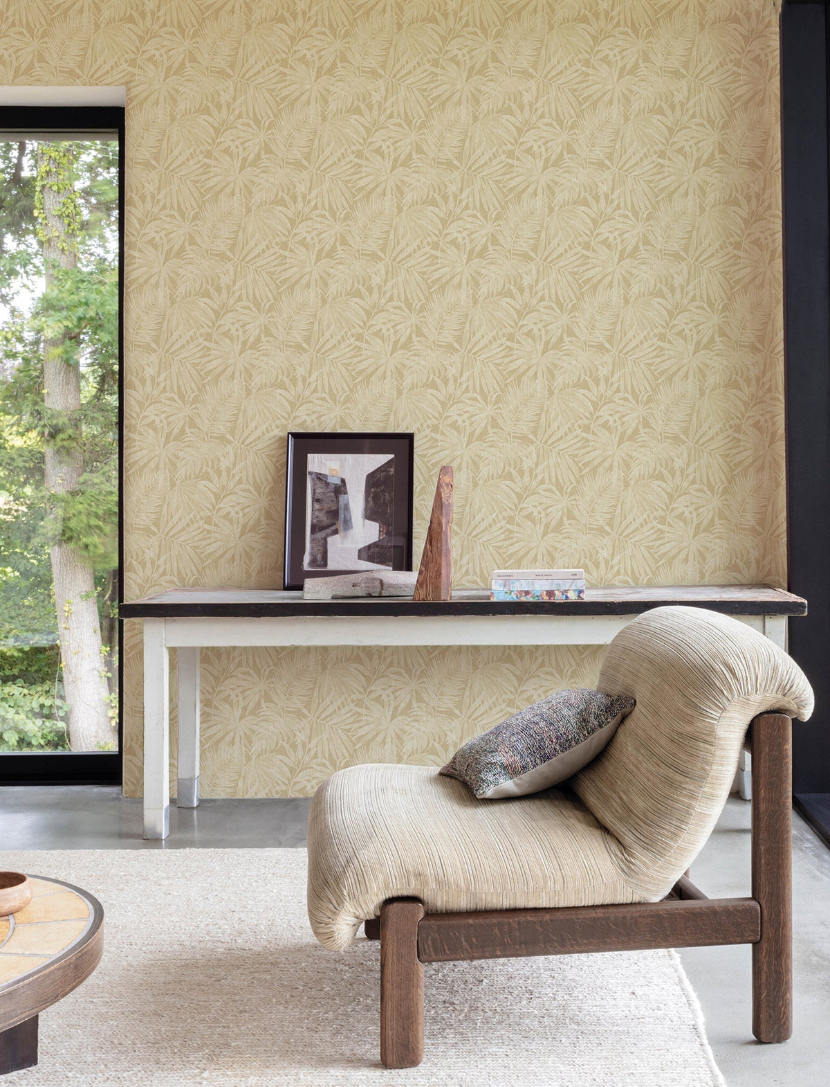 A cozy living room featuring Chesapeake Sonny Fronds Wallpaper by York Wallcoverings, with a beige textured chair, a patterned pillow, a slim table displaying framed art and a sculpture, and a large window with views of green trees outside.
