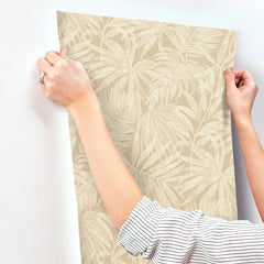 A person with a striped sleeve holds up Chesapeake Sonny Fronds Wallpaper by York Wallcoverings—featuring a beige, leaf-patterned design—against a white wall, preparing to apply it.