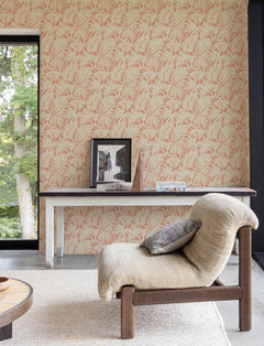 A cozy beige chair sits on a light rug near a table displaying framed photos, a book, and a decorative piece. The wall is adorned with York Wallcoverings’ Chesapeake Sonny Fronds Wallpaper in a pink palm leaf pattern, while large windows reveal green trees outside.