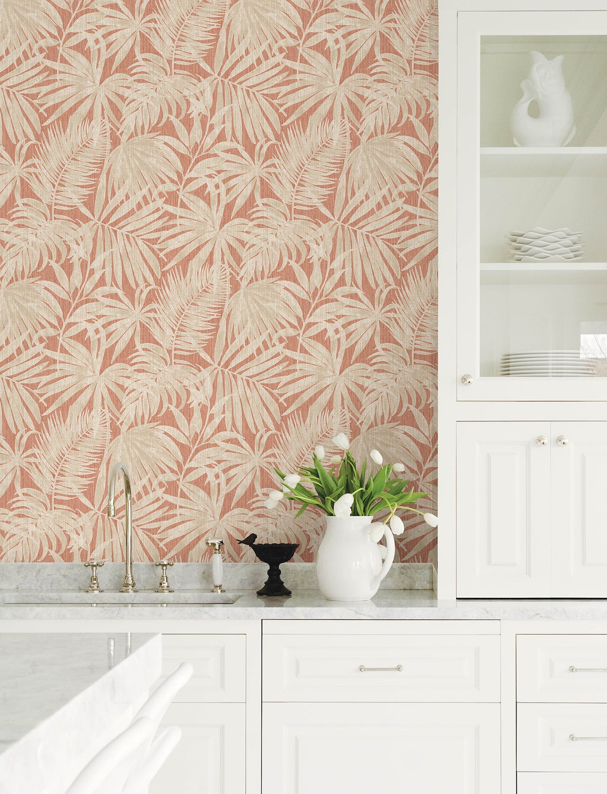 A white kitchen with a marble countertop, a sink, a white pitcher holding tulips, a black bird figurine, and a cabinet with glass doors. The wall is adorned with York Wallcoverings’ Chesapeake Sonny Fronds Wallpaper in pink and beige tropical leaf patterns.