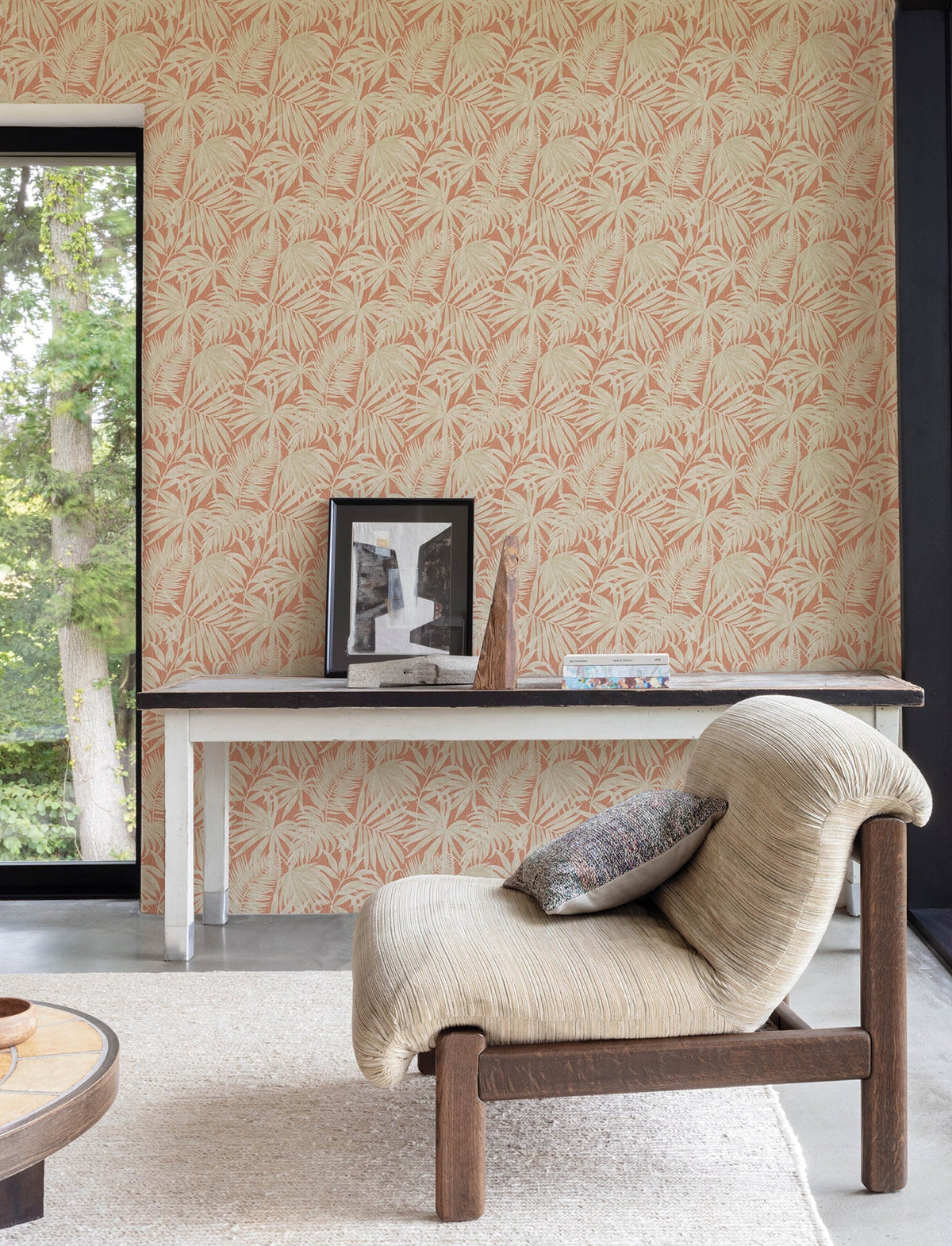Cozy room featuring a textured beige chair, a small table with framed photos, a geometric ornament, and books set against York Wallcoverings’ Chesapeake Sonny Fronds Wallpaper. A large window reveals green trees outside.