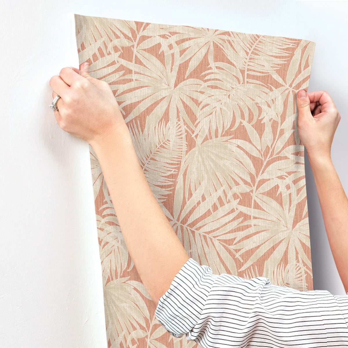 A person in a striped shirt holds up the Chesapeake Sonny Fronds Wallpaper by York Wallcoverings, featuring a beige tropical leaf pattern, against a white wall as they prepare to hang it.